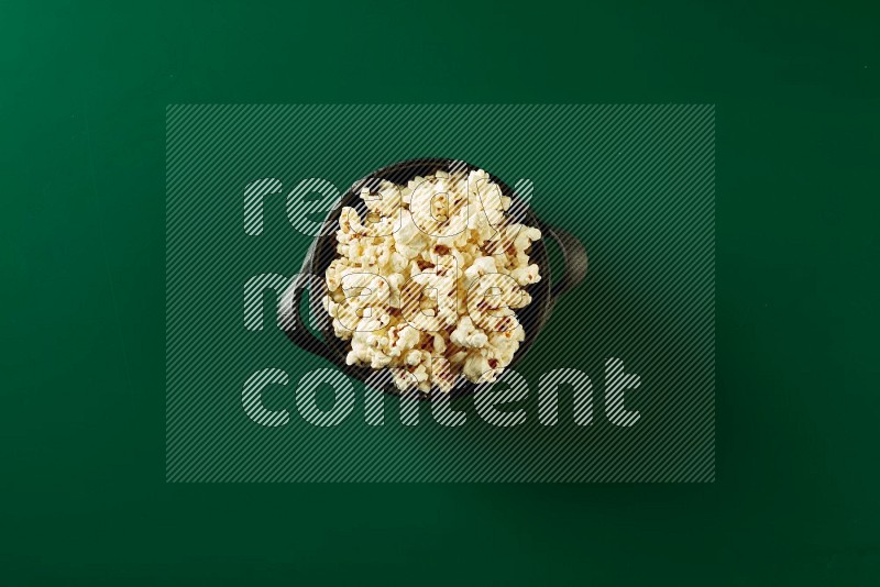A black ceramic bowl full of popcorn on a green background in a top view shot