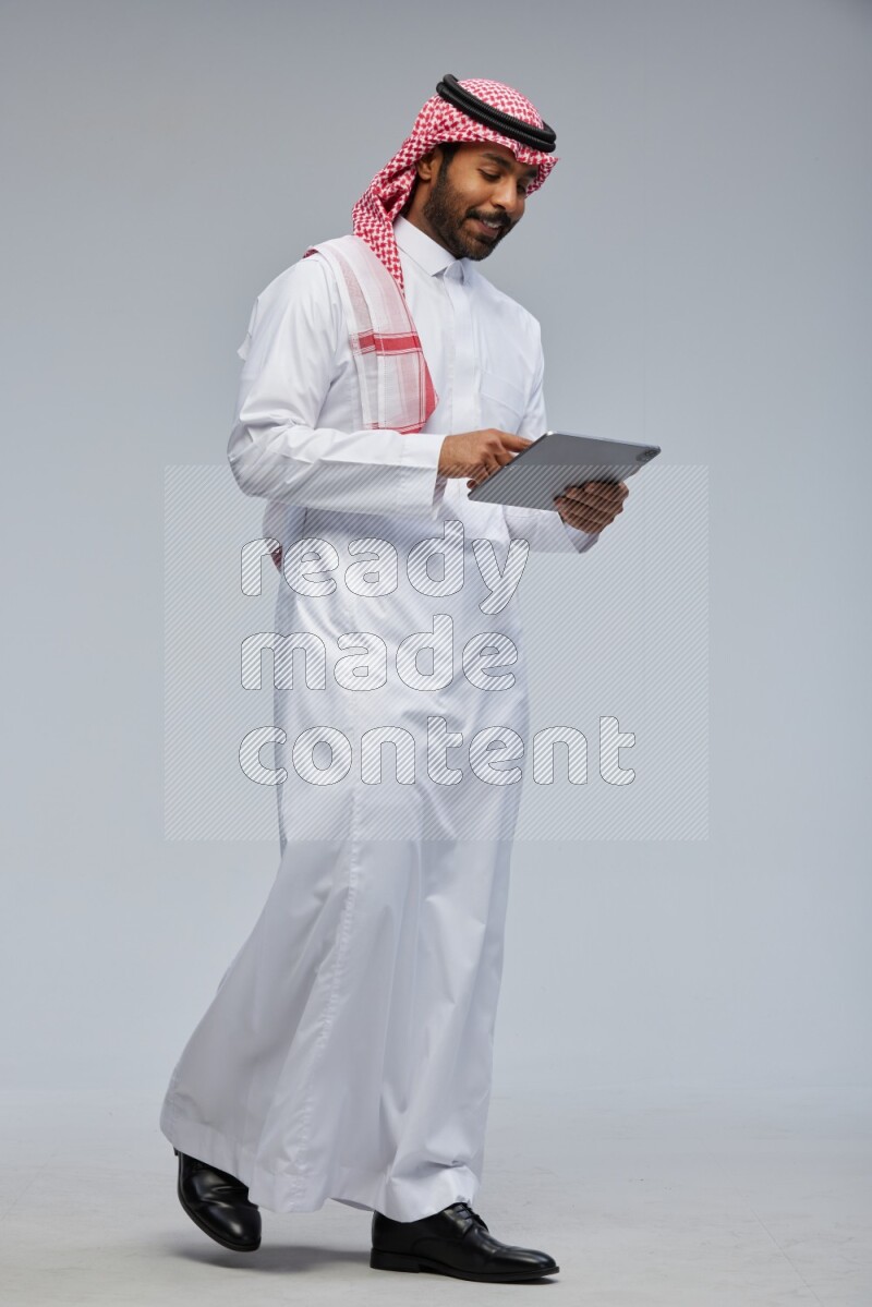 Saudi man Wearing Thob and shomag standing working on tablet on Gray background