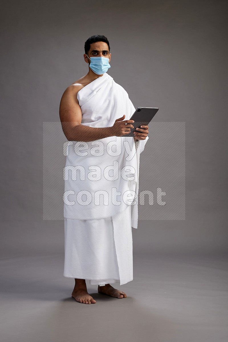 A man wearing Ehram with face mask Standing holding a tablet on gray background