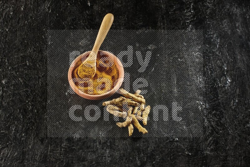 A wooden bowl and a wooden spoon full of turmeric powder with dried turmeric fingers on textured black flooring