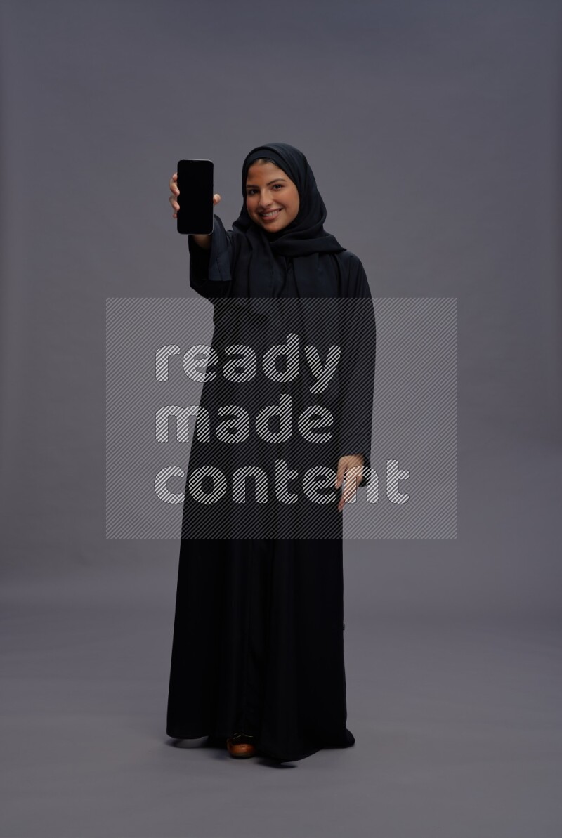Saudi woman wearing Abaya standing showing phone to camera on gray background