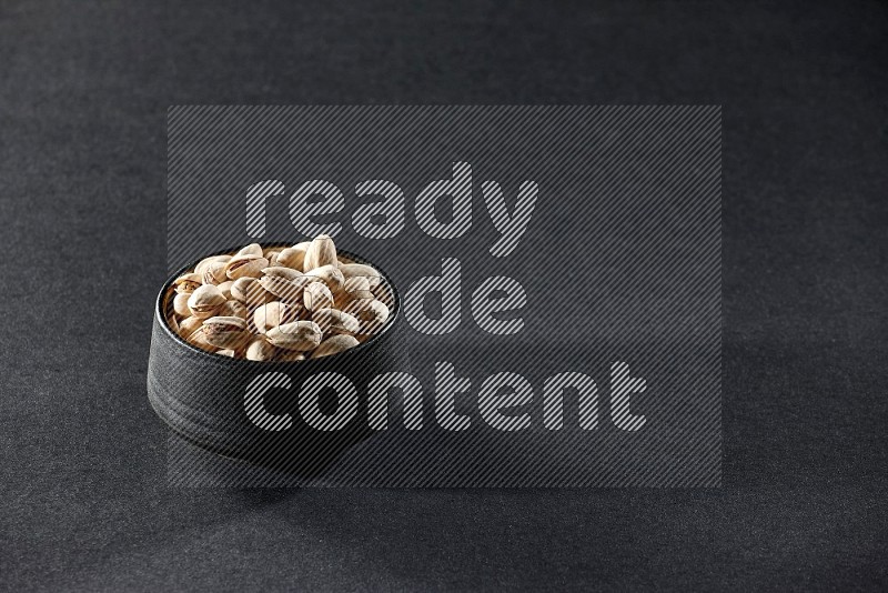 A black pottery bowl full of pistachios on a black background in different angles