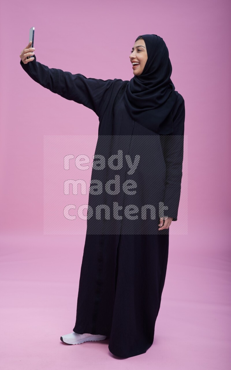 Saudi woman wearing Abaya standing taking selfie on pink background