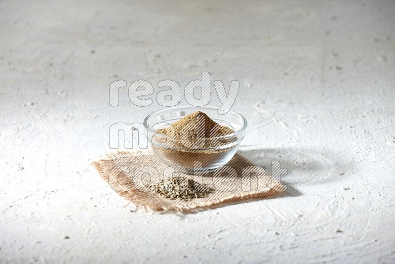 A glass bowl full of cumin powder with some of cumin seeds on burlap piece on a textured white flooring