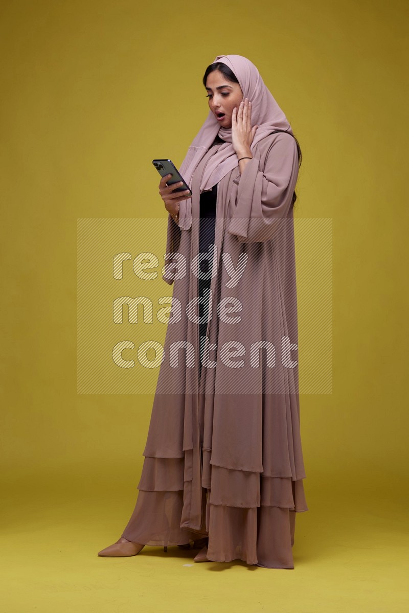 A woman Texting on her phone on a Yellow Background wearing Brown Abaya with Hijab