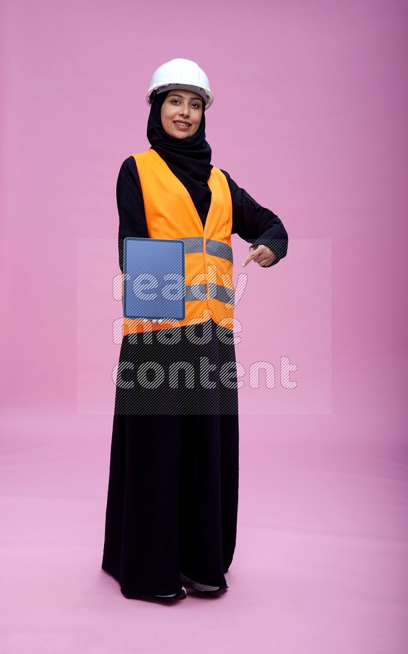 Saudi woman wearing Abaya with engineer vest and helmet standing showing tablet to camera on pink background