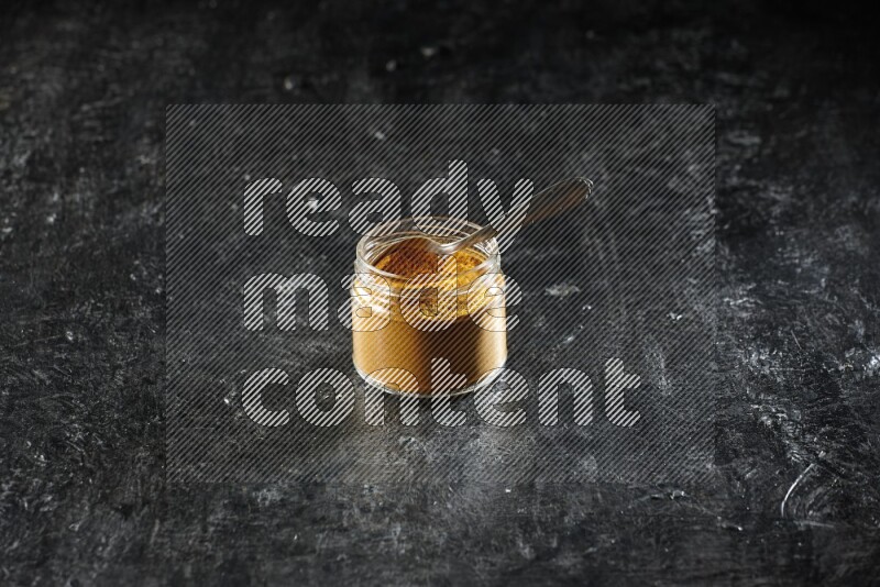 A glass jar and a metal spoon full of turmeric powder on a textured black flooring