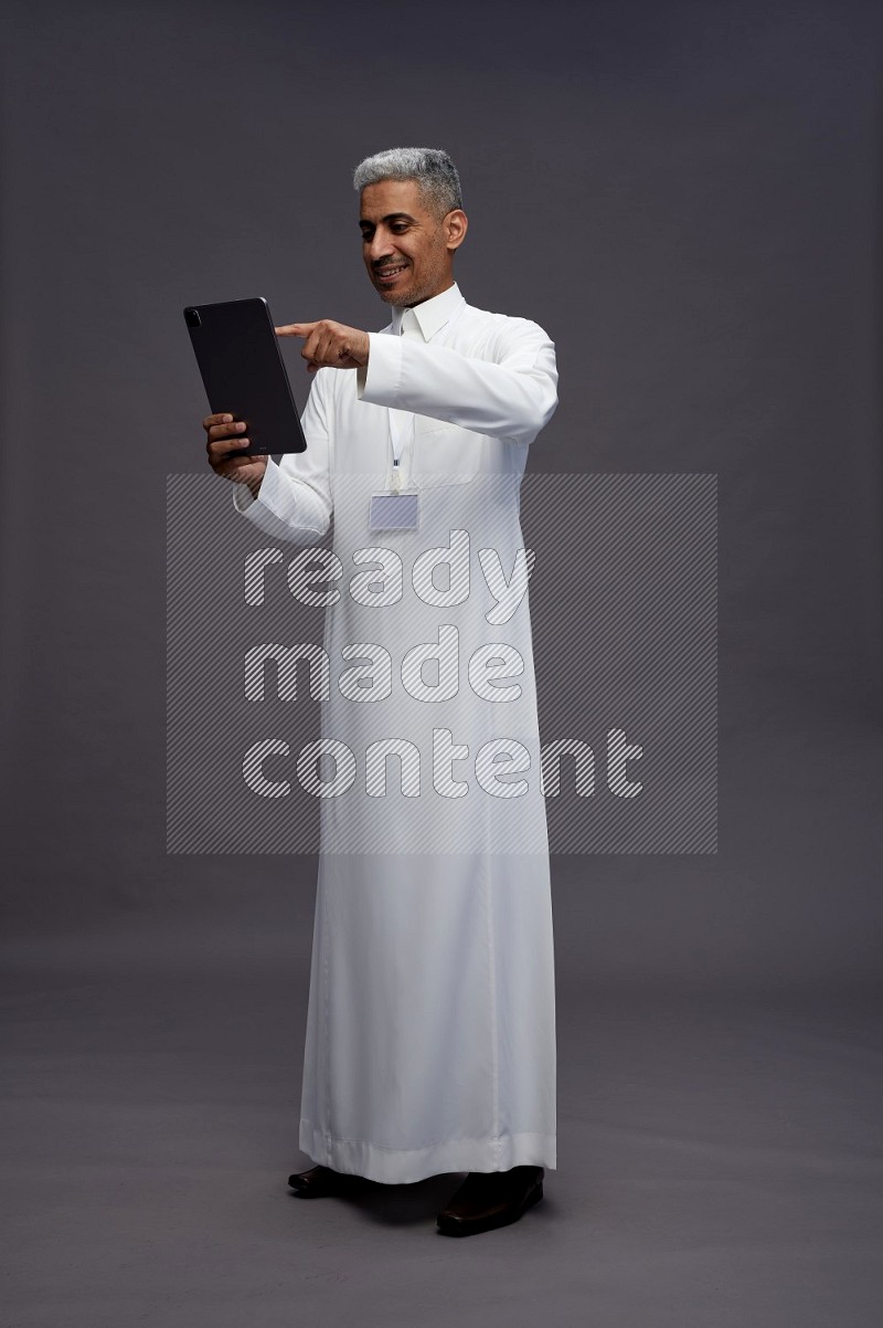 Saudi man wearing thob with neck strap employee badge standing working on tablet on gray background