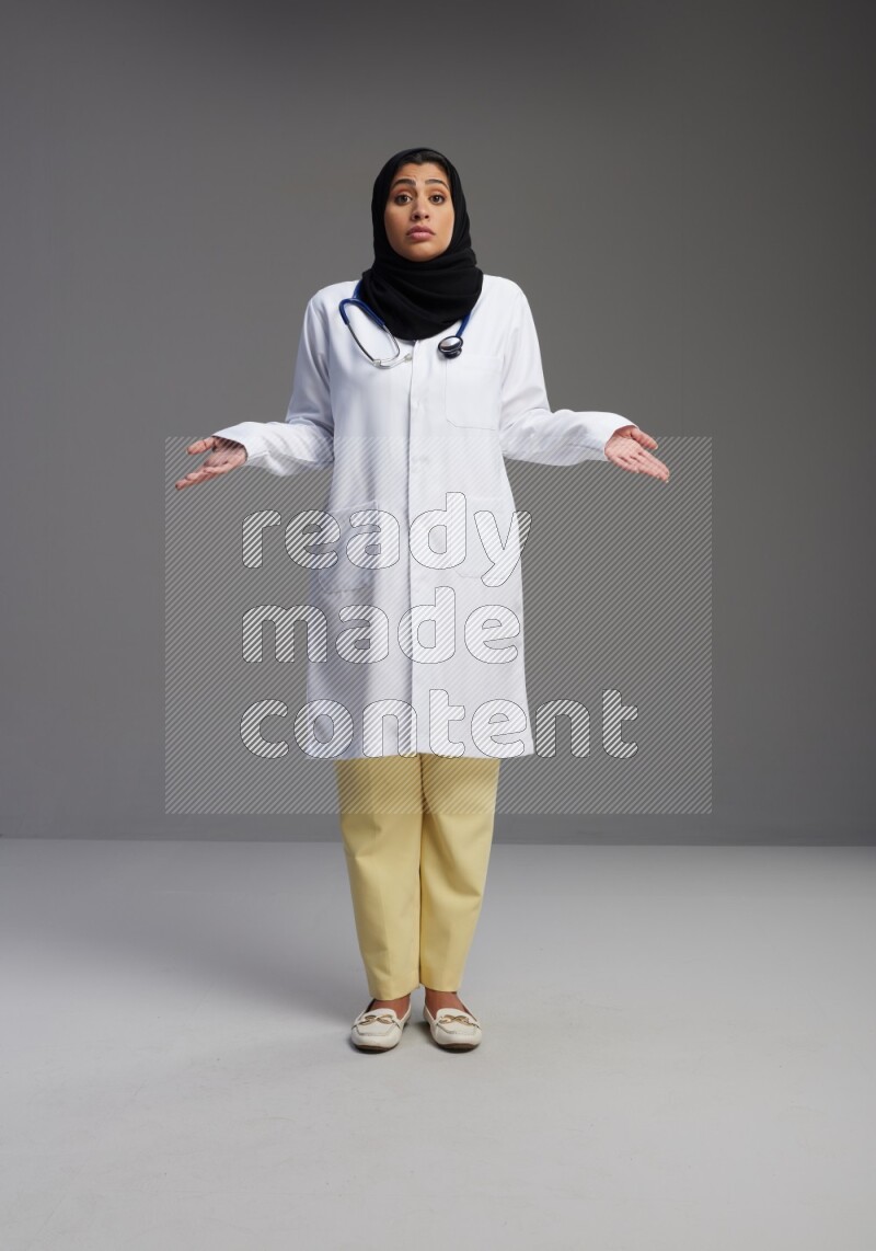 Saudi woman wearing lab coat with stethoscope standing interacting with the camera on Gray background