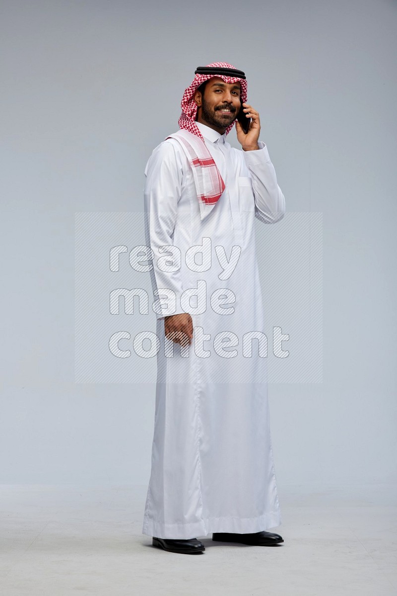 Saudi man Wearing Thob and shomag standing talking on phone on Gray background