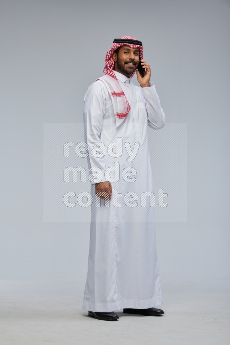 Saudi man Wearing Thob and shomag standing talking on phone on Gray background
