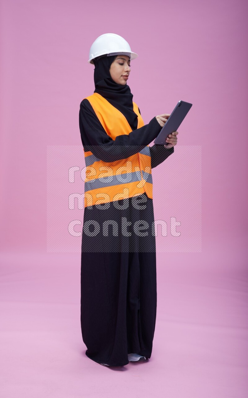 Saudi woman wearing Abaya with engineer vest and helmet standing working on tablet on pink background