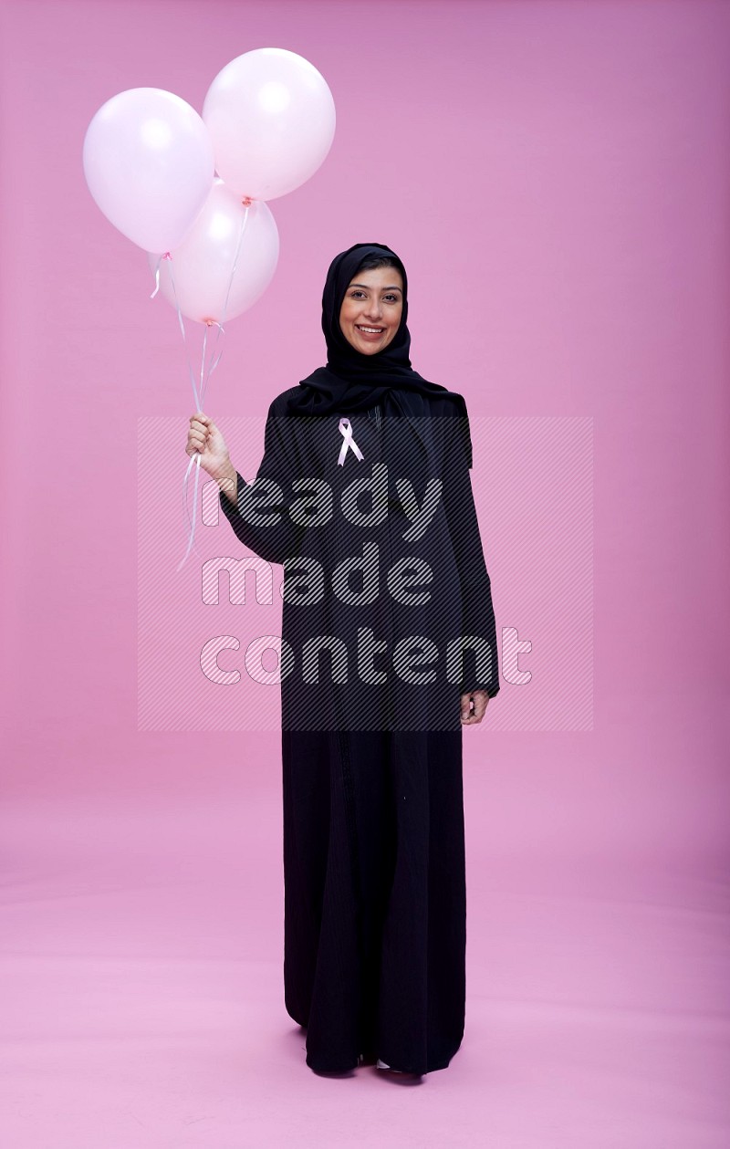 Saudi woman wearing pink ribbon on Abaya standing holding balloon on plain pink background