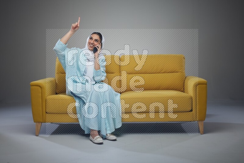 A Saudi woman wearing a light blue Abaya and a white head scarf sitting on a yellow sofa and talking in the phone while making different poses eye level on a grey background