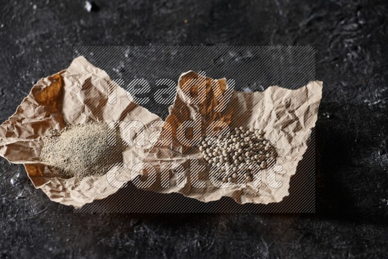 White pepper beads and powder in 2 crumpled paper on textured black flooring
