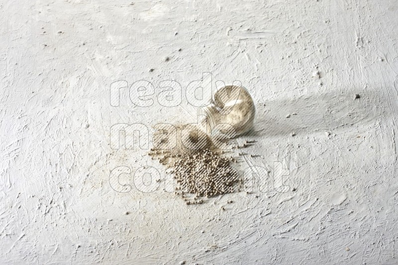 A flipped herbal glass jar full of white pepper powder with spilled powder and beads on textured white flooring
