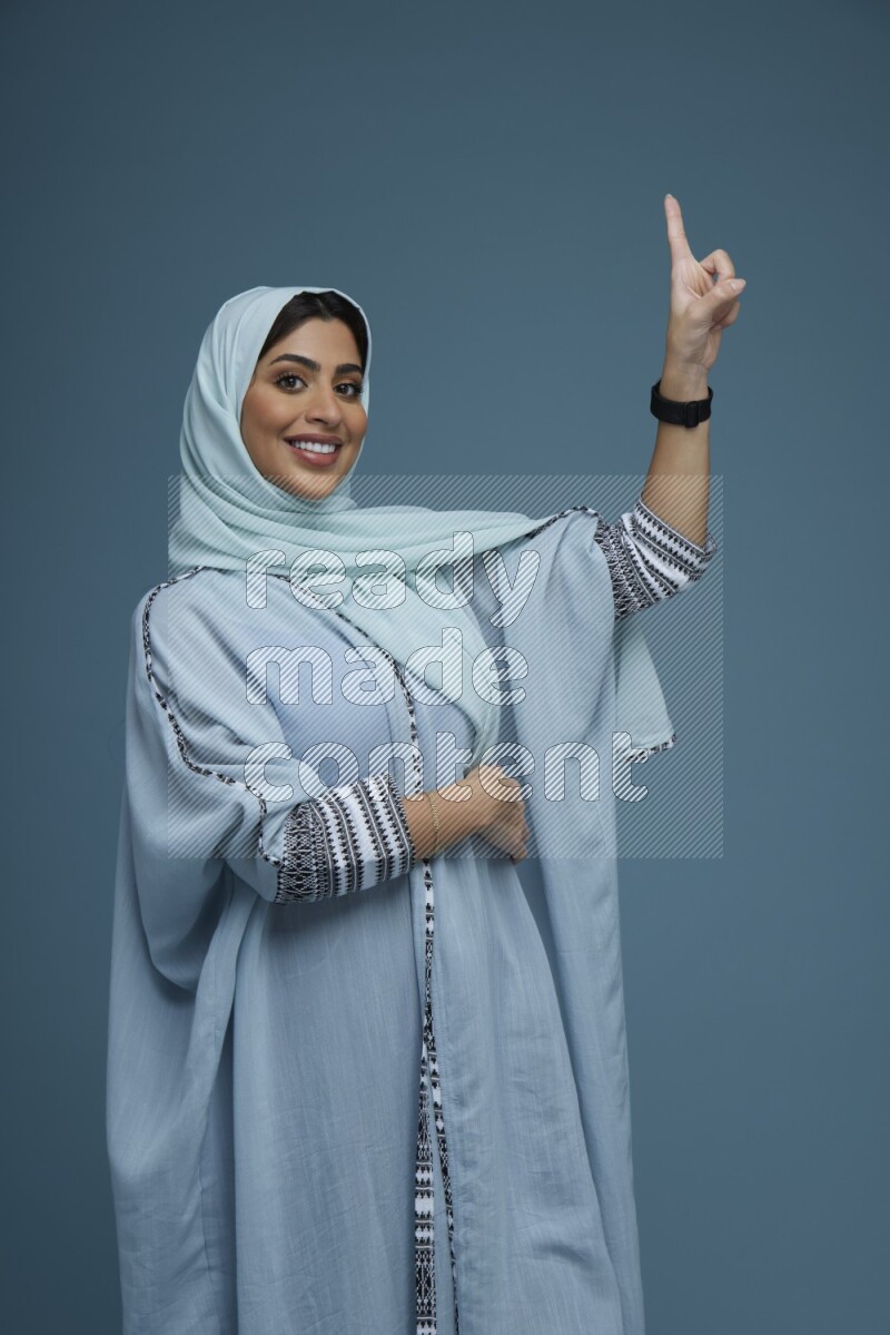 A Saudi woman pointing in a blue background wearing a blue Abaya with hijab