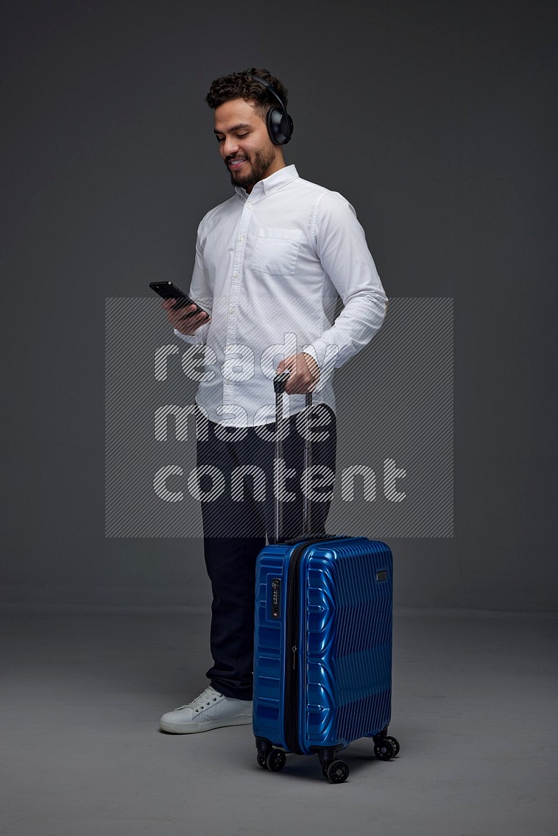 A man wearing smart casual and headphone holding luggage eye level on a gray background
