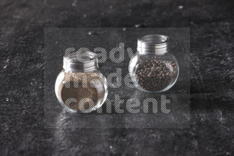 2 glass spice jars full of black pepper powder and black pepper beads on textured black flooring
