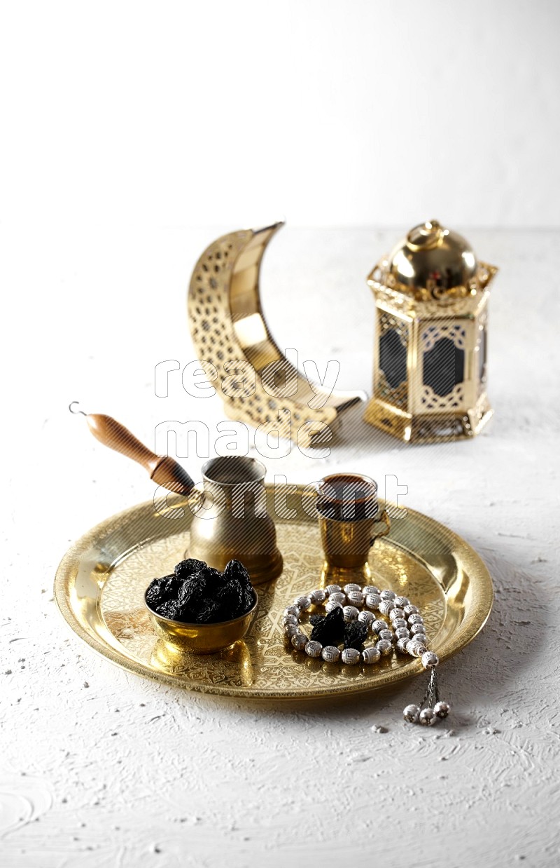 Dried plums in a metal bowl with coffee and prayer beads on a tray beside lanterns in a light setup