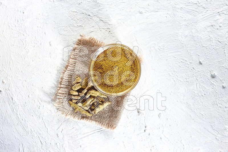 A glass bowl full of turmeric powder and dried turmeric whole finger on a piece of burlap on a textured white flooring