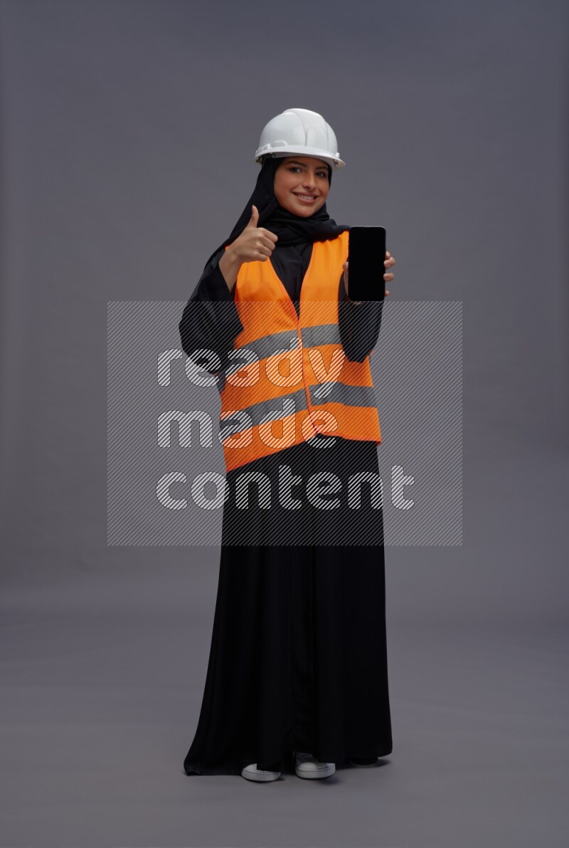 Saudi woman wearing Abaya with engineer vest standing showing phone to camera on gray background