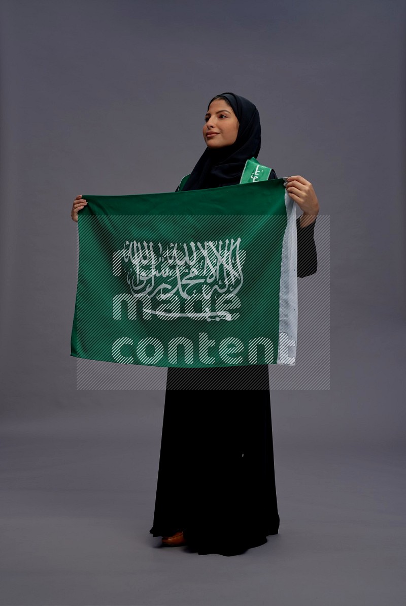 Saudi woman wearing Abaya standing holding Saudi flag on gray background