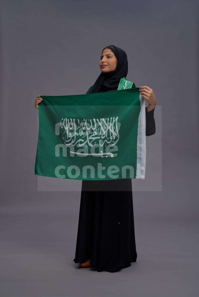 Saudi woman wearing Abaya standing holding Saudi flag on gray background