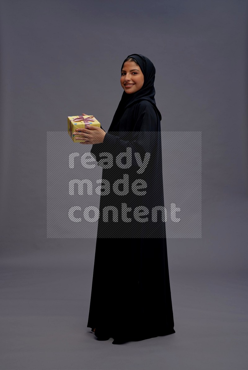 Saudi woman wearing Abaya standing holding gift box on gray background