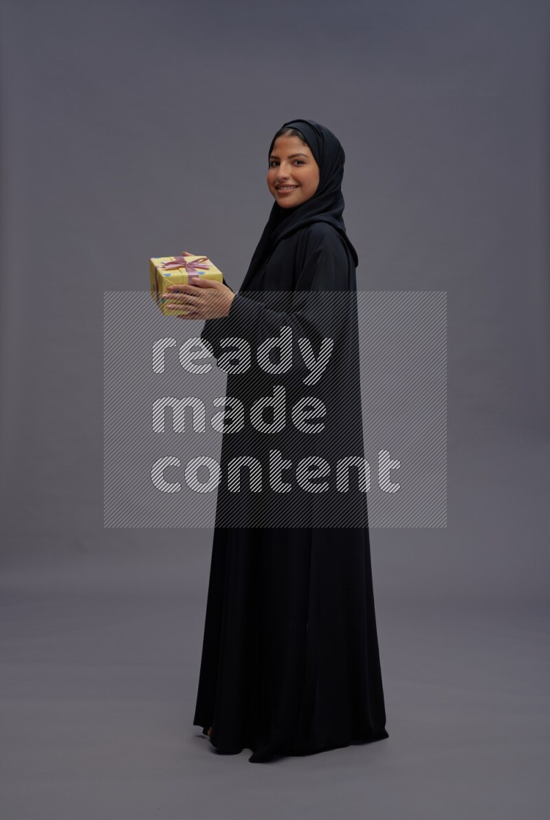 Saudi woman wearing Abaya standing holding gift box on gray background