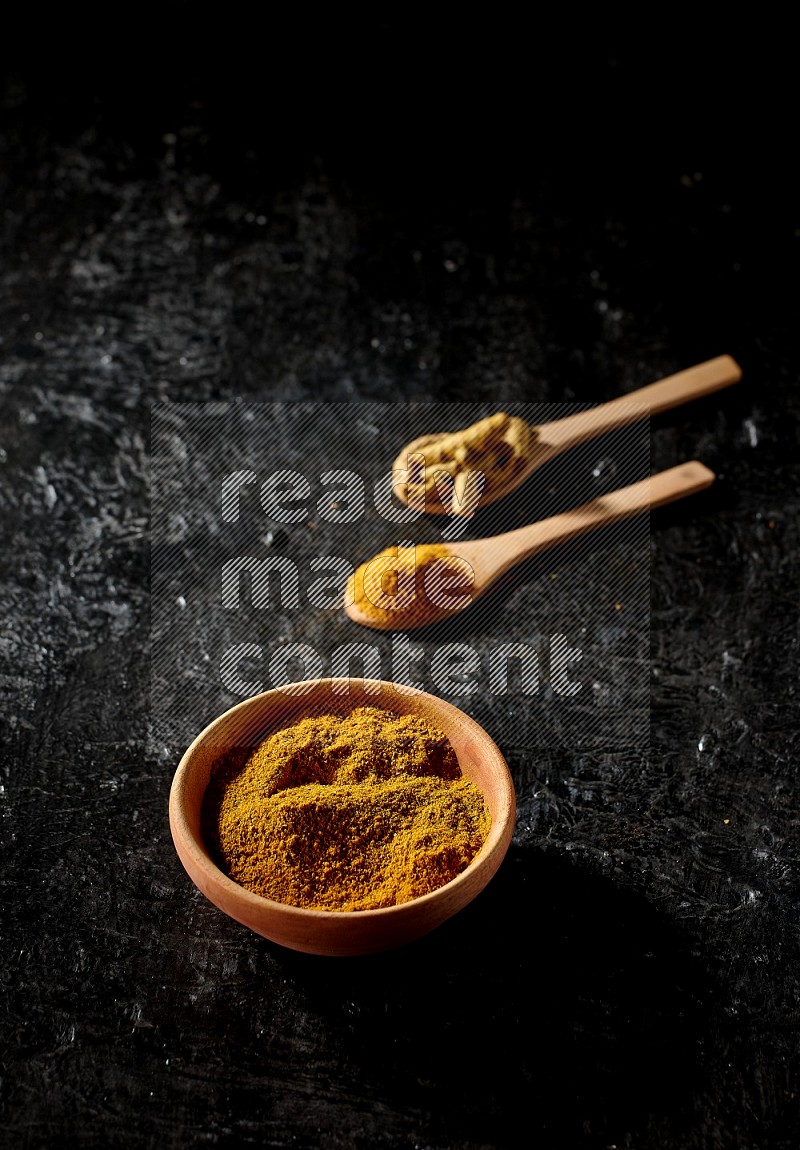 A wooden bowl full of turmeric powder and 2 wooden spoons full of dried turmeric finger and turmeric powder on textured black flooring