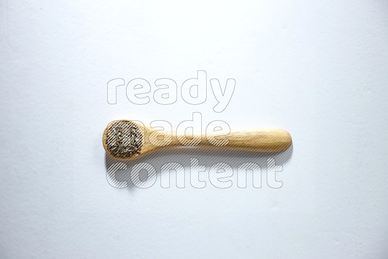 A wooden spoon full of cumin seeds on a white flooring