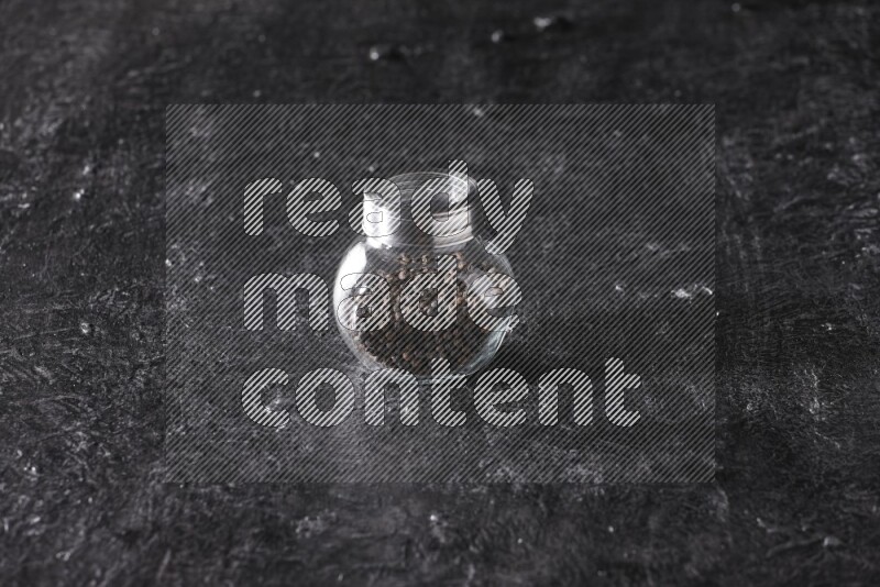 A glass spice jar full of black pepper beads on textured black flooring