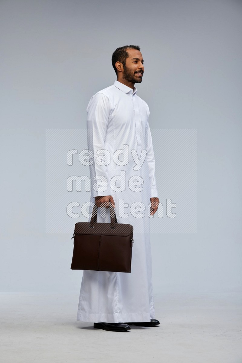 Saudi man wearing thob standing holding suitcase on gray background