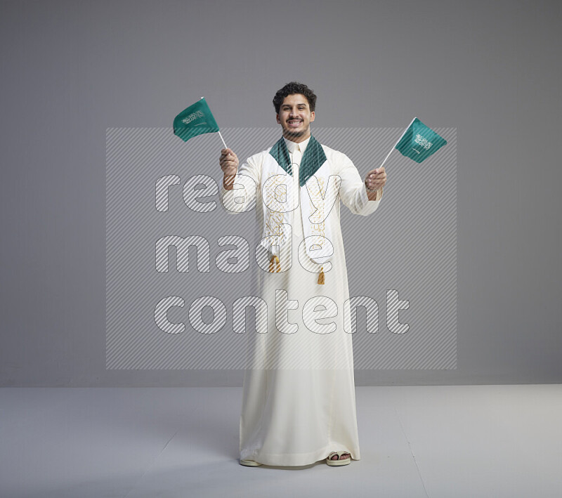 A Saudi man standing wearing thob and saudi flag scarf and holding small saudi flag on gray background