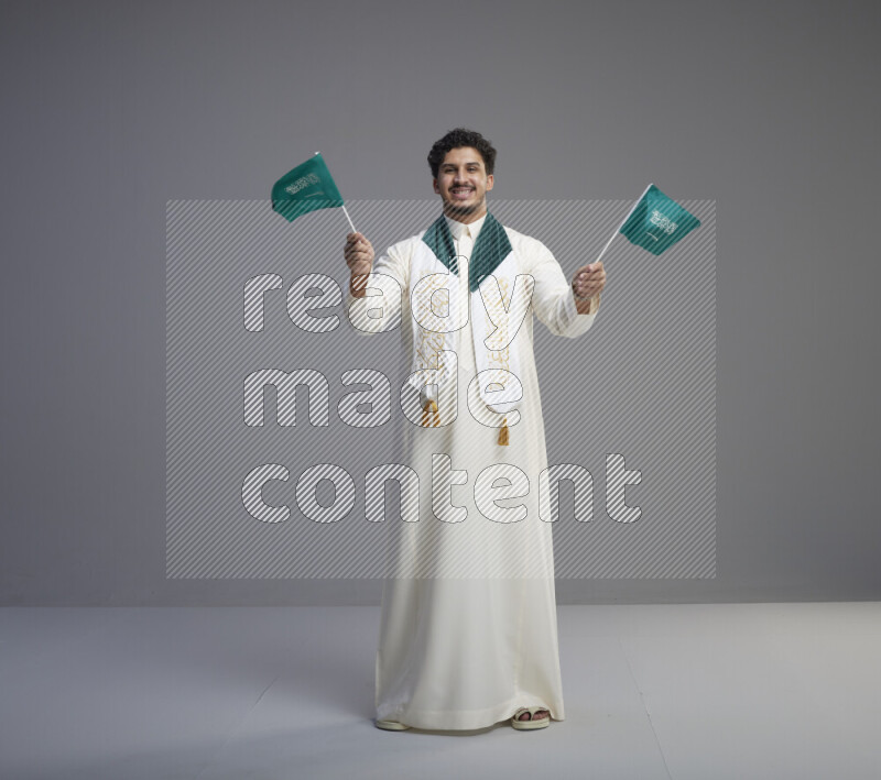 A Saudi man standing wearing thob and saudi flag scarf and holding small saudi flag on gray background
