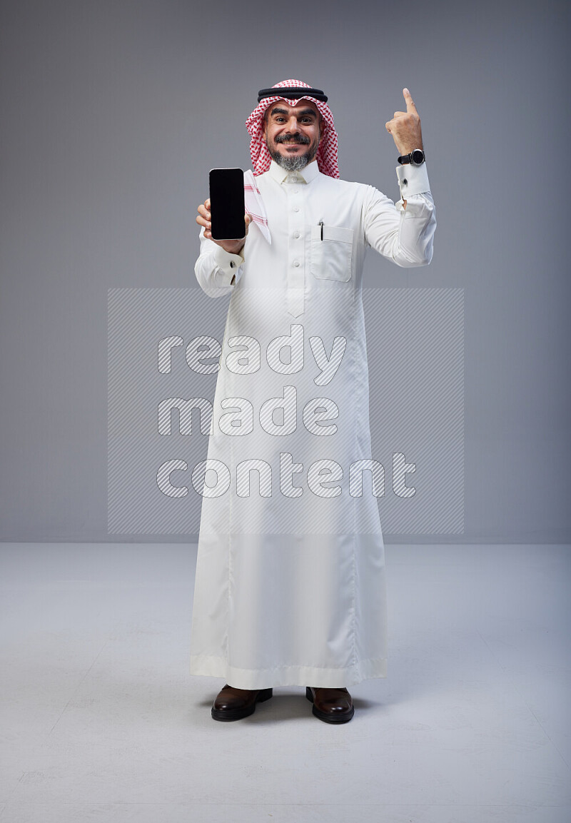 Saudi man Wearing Thob and red Shomag standing showing phone to camera on Gray background