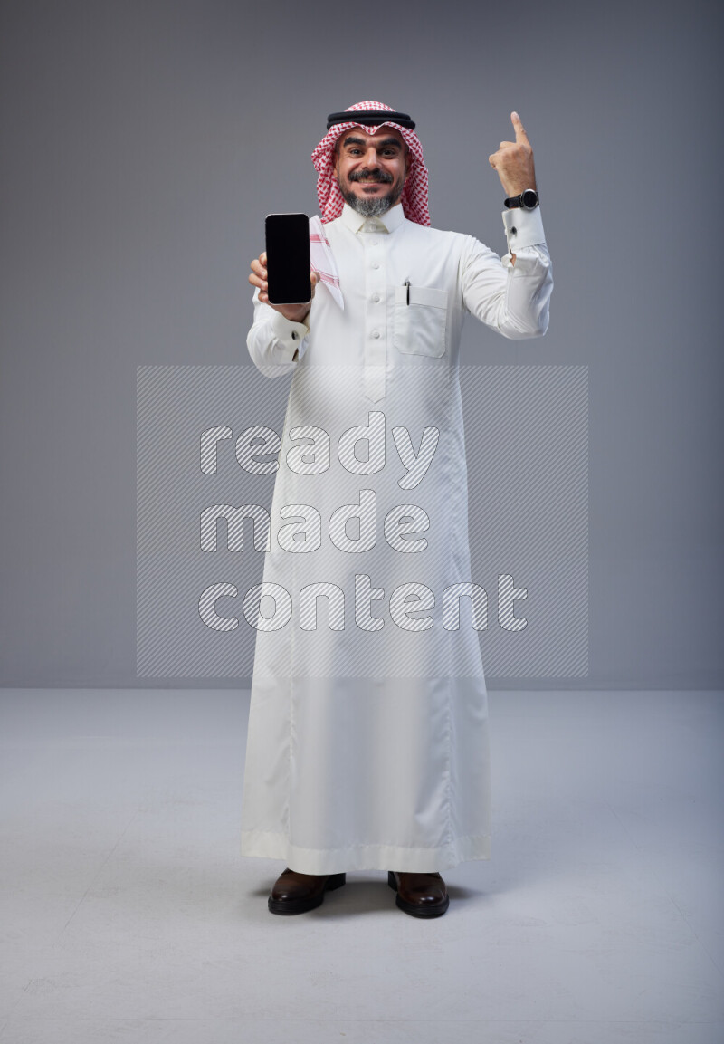 Saudi man Wearing Thob and red Shomag standing showing phone to camera on Gray background