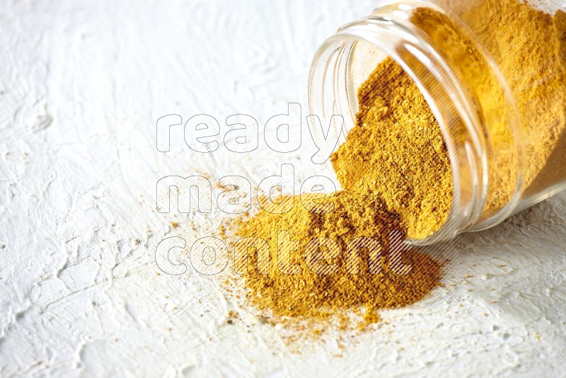 A flipped glass spice jar full of turmeric powder and powder fell out it on textured white flooring