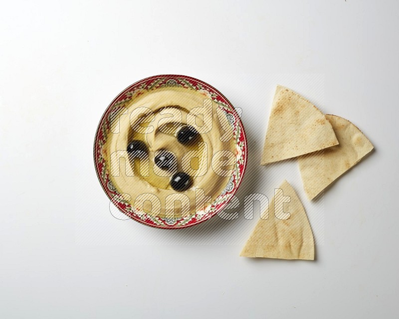 Hummus in a red plate with patterns garnished with black olives on a white background