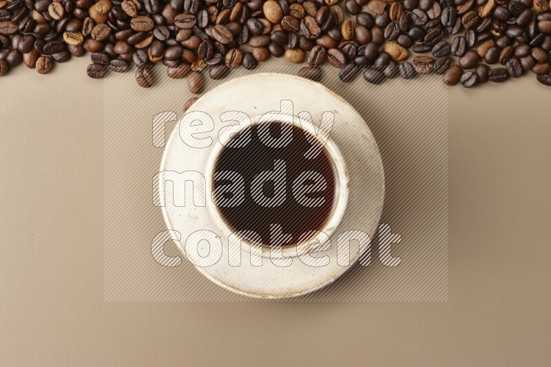A beige pottery cup of coffee surrounded by roasted coffee beans on beige background