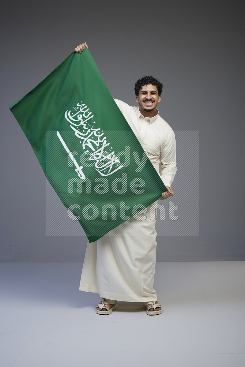 A saudi man standing wearing thob holding big saudi flag on gray background