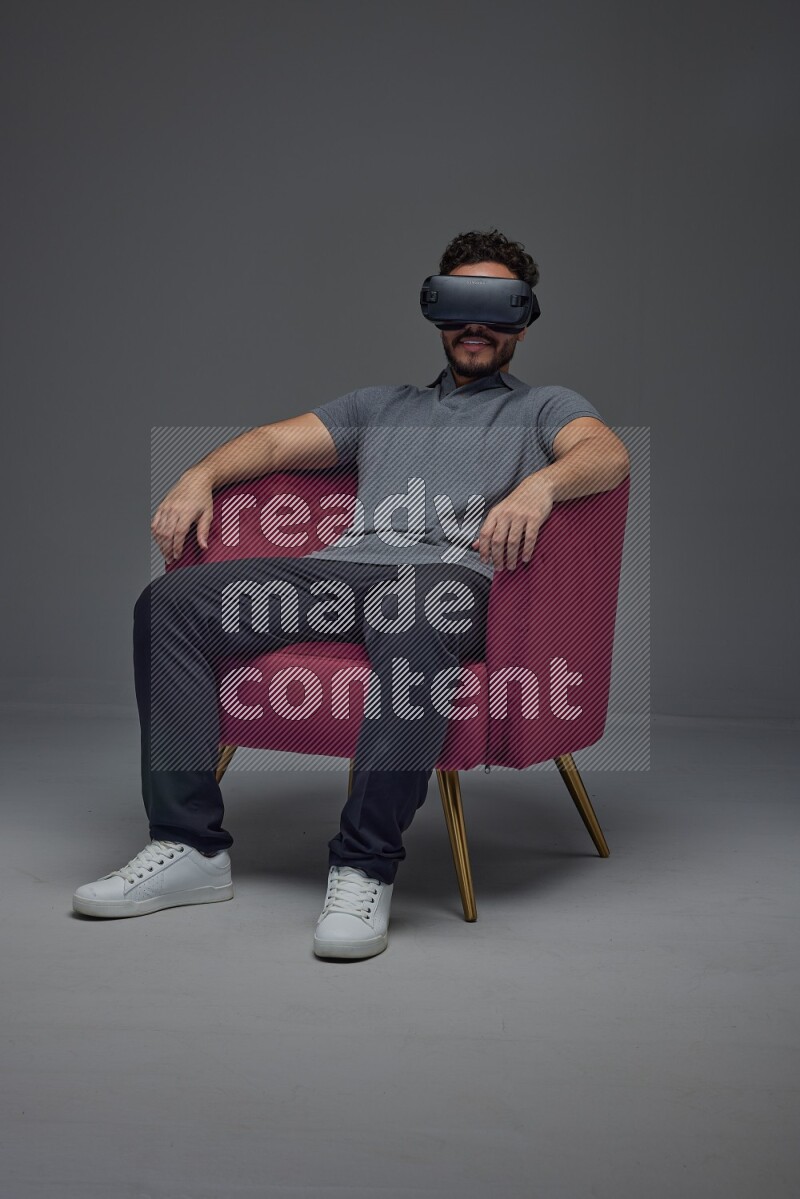 A man wearing casual and wearing VR while sitting on a burgundy chair eye level on a gray background