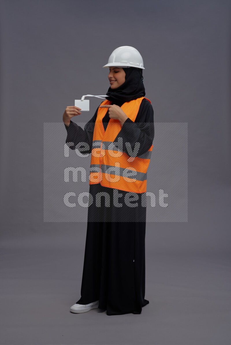 Saudi woman wearing Abaya with engineer vest with neck strap employee badge standing interacting with the camera on gray background
