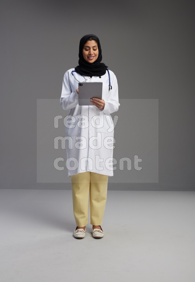 Saudi woman wearing lab coat with stethoscope standing working on tablet on Gray background
