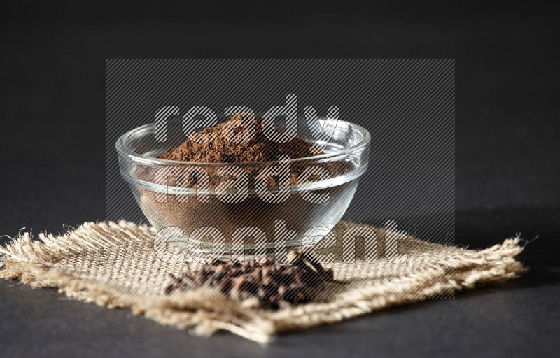 A glass bowl full of cloves powder with cloves grains on a burlap piece on a black flooring