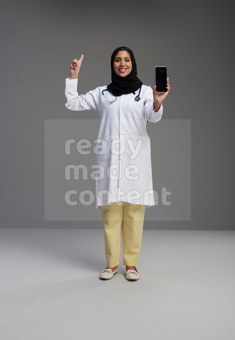 Saudi woman wearing lab coat with stethoscope standing showing phone to camera with sign in the back on Gray background