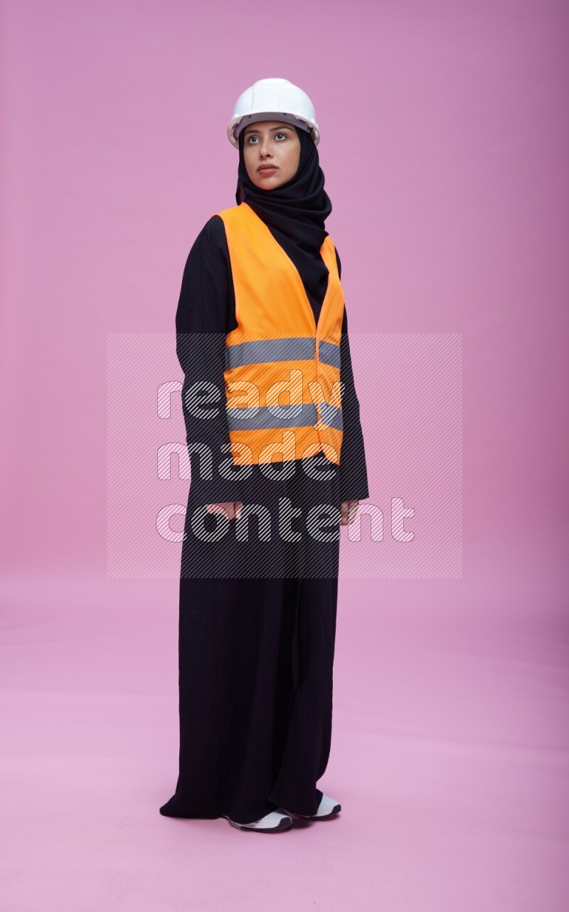 Saudi woman wearing Abaya with engineer vest and helmet standing interacting with the camera on pink background