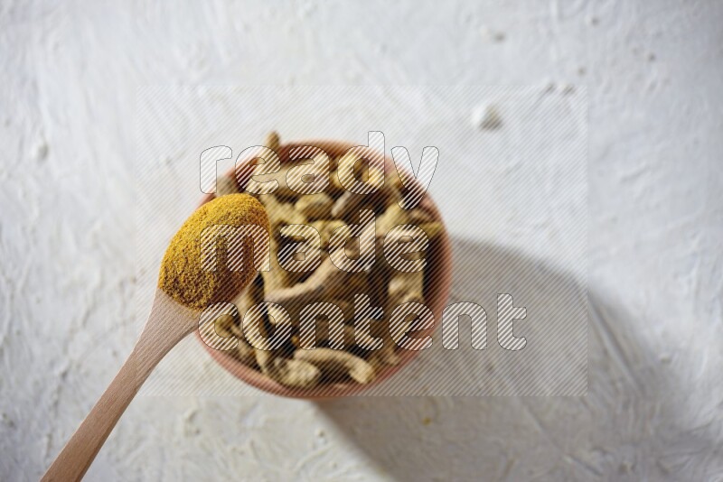 A wooden spoon full of turmeric powder above a wooden bowl full of dried turmeric whole fingers on a textured white flooring