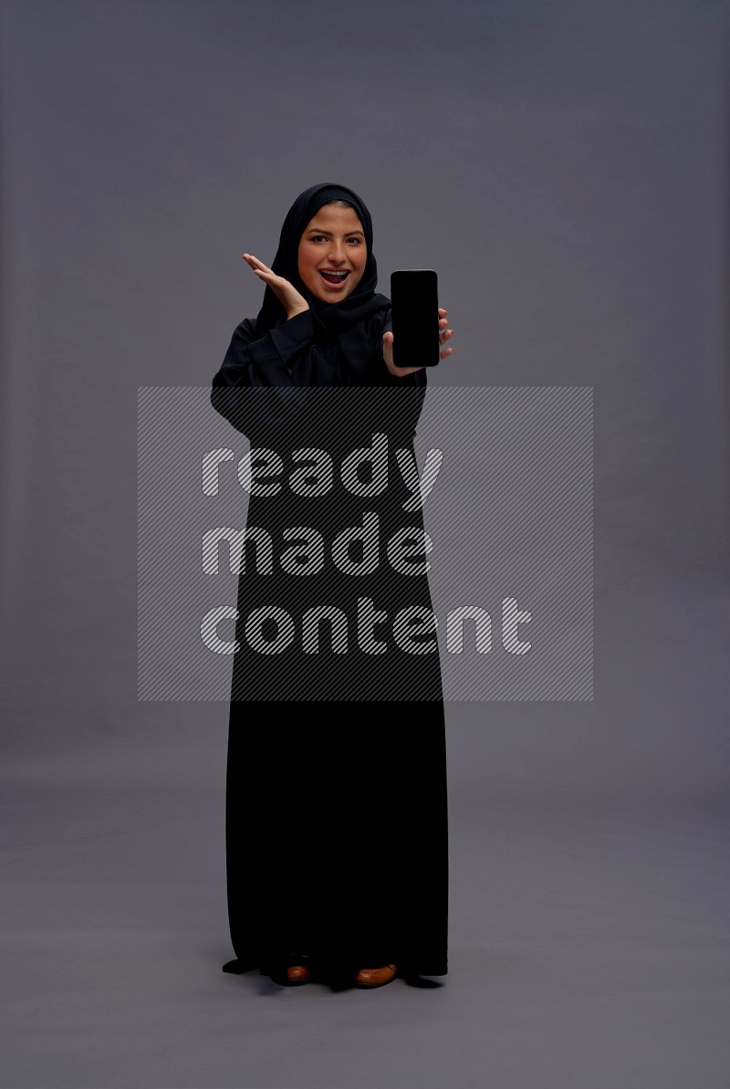 Saudi woman wearing Abaya standing showing phone to camera on gray background